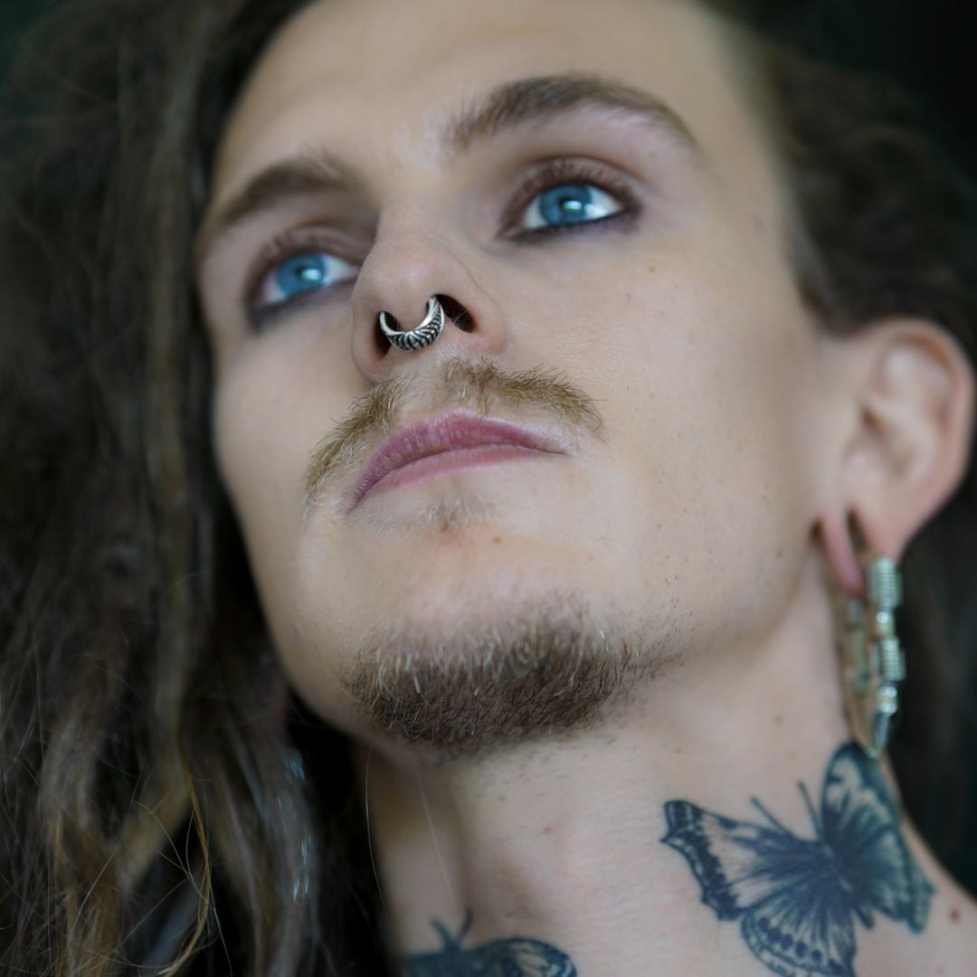 A person with light skin and blue eyes is looking upward. They have long, wavy hair, multiple ear piercings, and a sleek Darma Biomechanical Clicker Hoop Septum Nose Ring in Silver from Kaya Movement. Additionally, they display a butterfly tattoo on their neck and have eyeliner applied around their eyes, adding a touch of post-apocalyptic body jewelry flair.